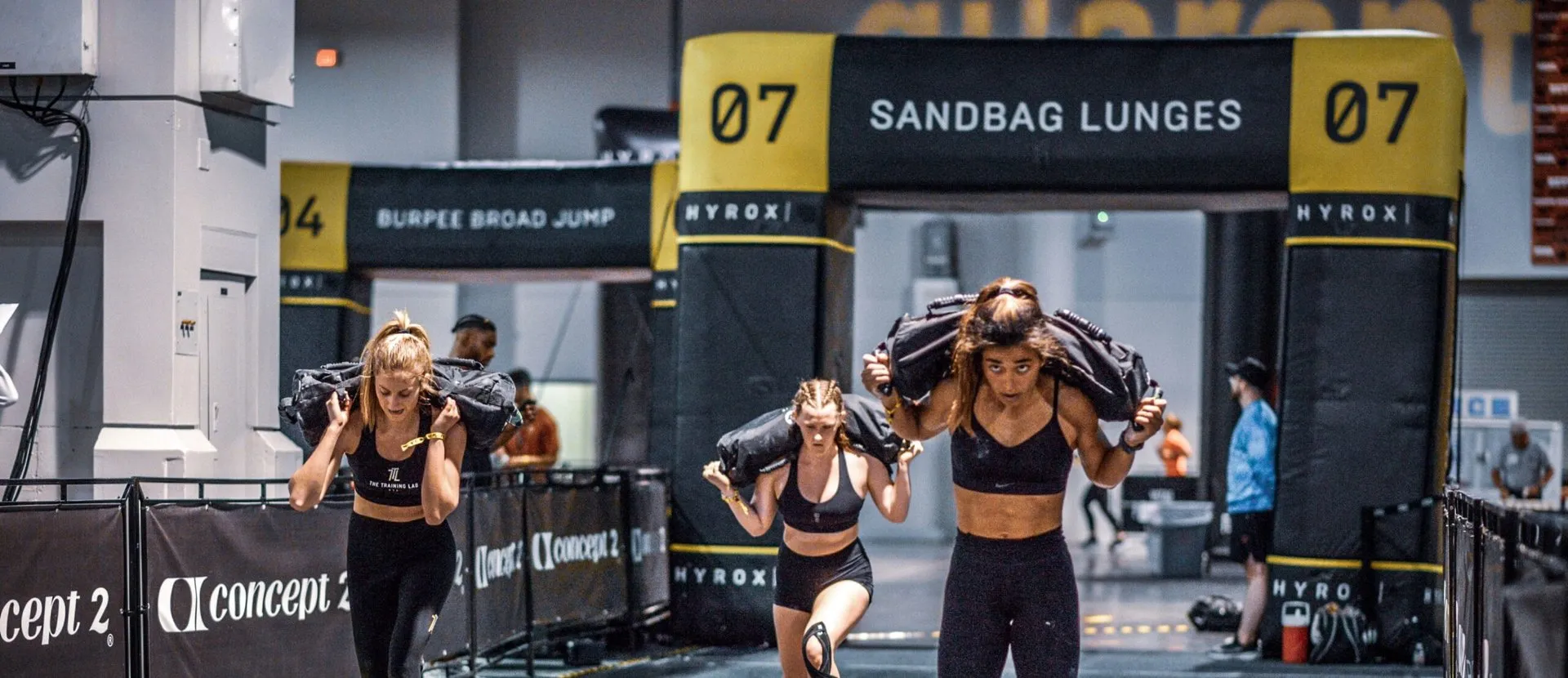 Tres atletas avanzan en zancadas con el saco a los hombros bajo el arco de la estación «07 Sandbag Lunges» de una carrera HYROX